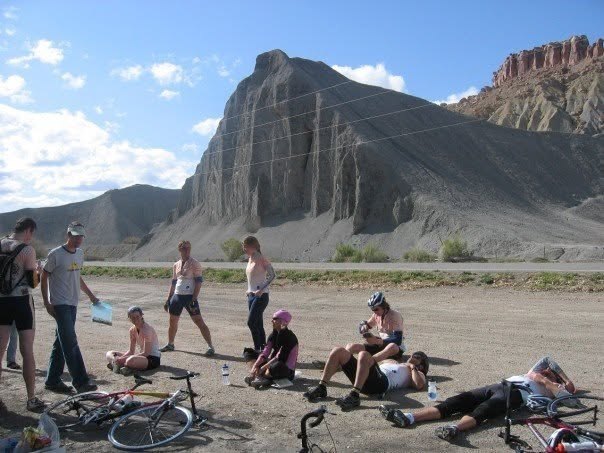 Riders and their bikes resting on the roadside below a grey rock butte and red cliffs