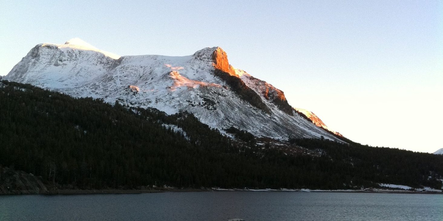 First light on a snow-dusted peak above a dark alpine lake, the forested shore still in shadow