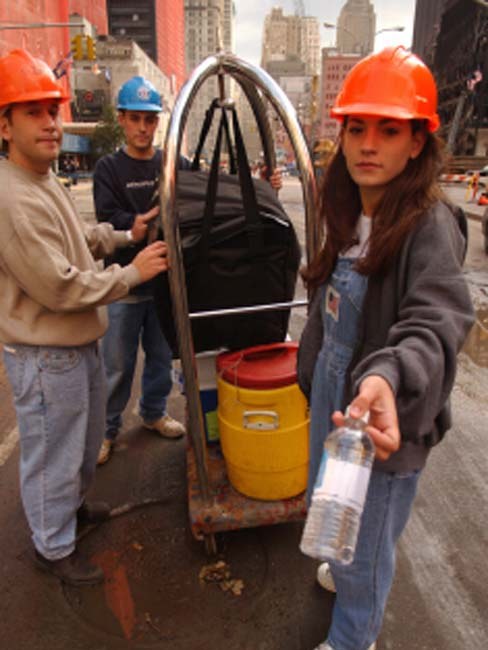 Three volunteers in hard hats with a cart of water coolers on a street near Ground Zero; a young woman in overalls holds out a bottle of water