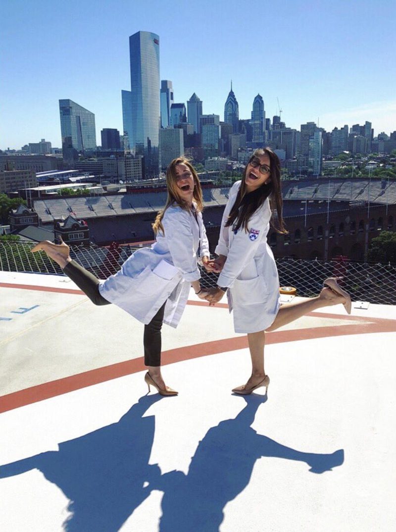 Two surgeons in white coats kicking up their heels on a rooftop above a city skyline