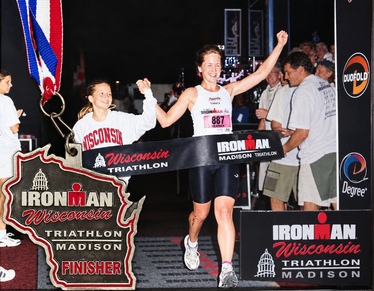 Heather, arms raised and smiling, breaking the finish-line tape at Ironman Wisconsin in Madison, holding hands with a girl in a Wisconsin sweatshirt who runs beside her; the race&rsquo;s finisher medal hangs in the foreground.
