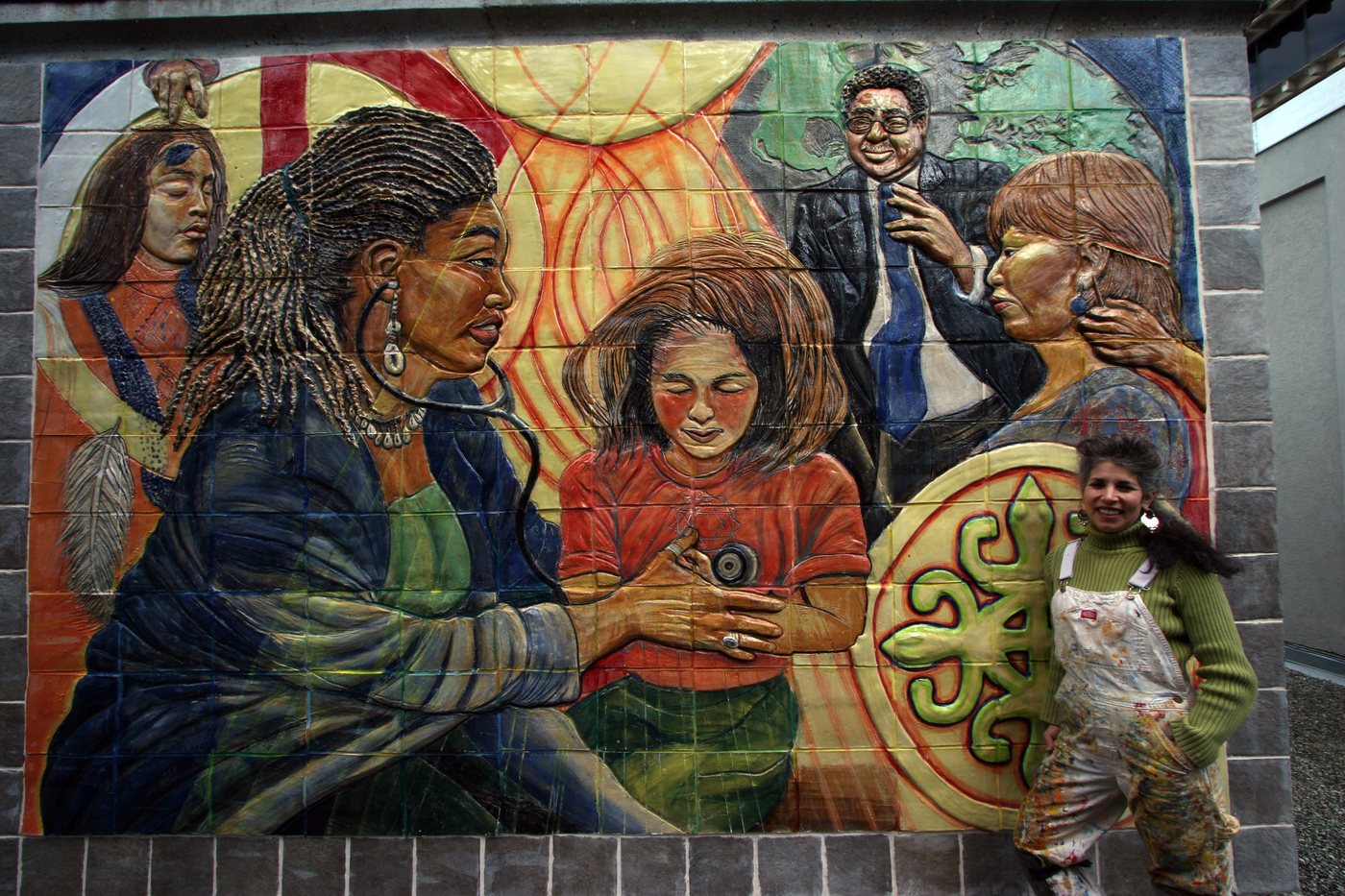 Juana Alicia, smiling in paint-spattered overalls and a green sweater, stands beside her ceramic bas-relief mural Traditions at UCSF: a woman with a stethoscope listens to a girl&rsquo;s heart, surrounded by a woman receiving a blessing, a man in a suit, a woman whose neck is being examined, and a large green emblem.