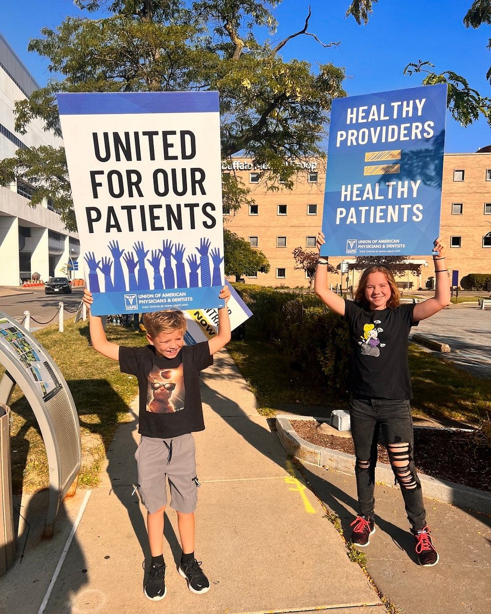 Two children on a sidewalk outside a hospital, each holding a Union of American Physicians and Dentists sign above their heads. One reads &ldquo;United for our patients,&rdquo; the other &ldquo;Healthy providers = healthy patients.&rdquo;