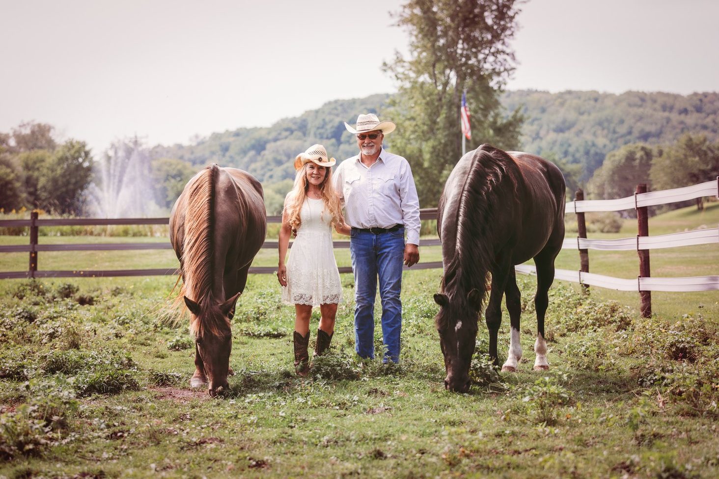 John and Jill Logghe standing between two dark horses in a fenced pasture, both in straw cowboy hats; Jill in a white lace dress and boots, John in a white western shirt and jeans, with wooded hills and an American flag behind them