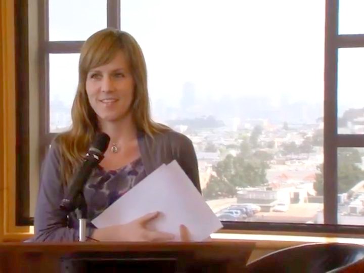 Heather speaking at a podium, the San Francisco skyline behind her